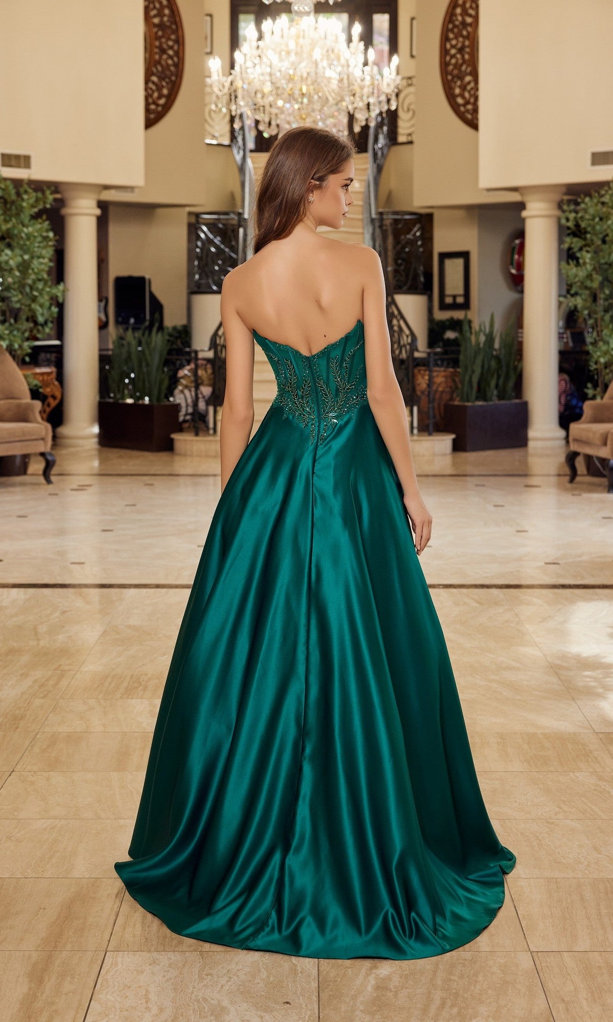 A woman wearing the Nox Anabel Y1798 long prom dress, featuring a strapless satin corset bodice and elegant A-line silhouette, stands in an opulent, chandelier-lit lobby with her back to the camera.